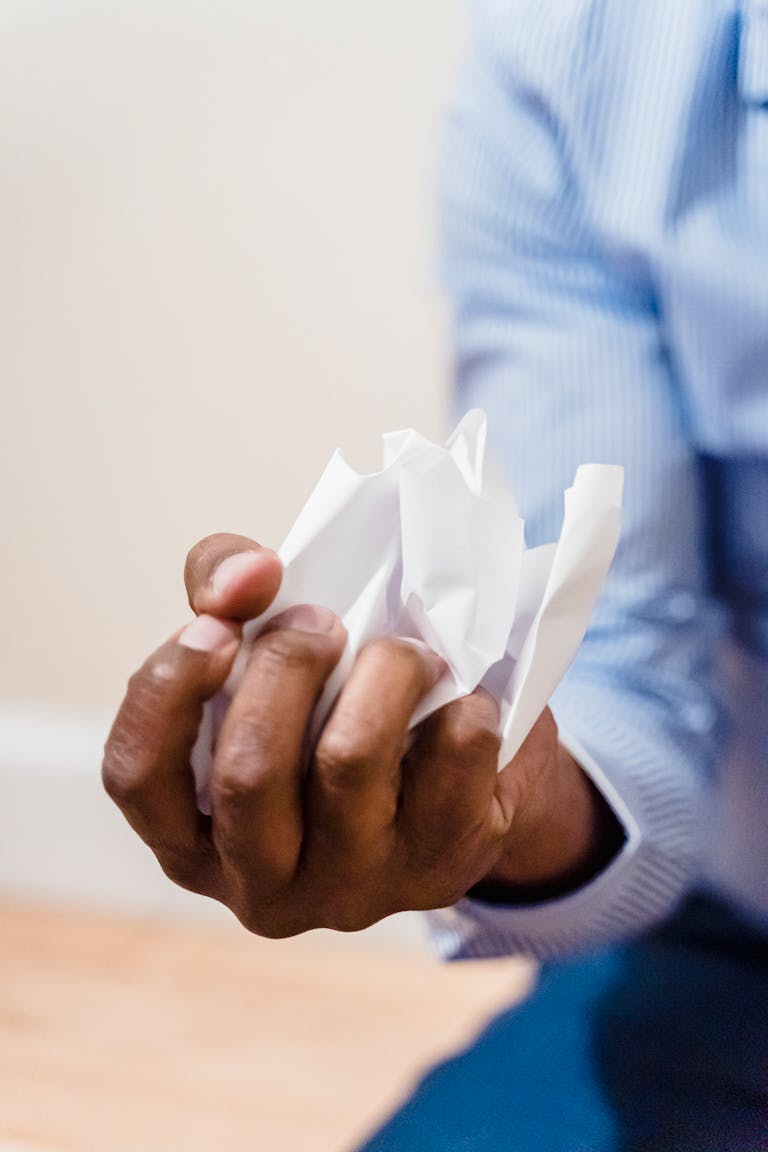 A hand in a blue sleeve tightly grips a crumpled piece of white paper indoors.